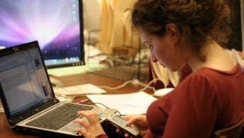 A girl navigates her phone while using her laptop while studying at home